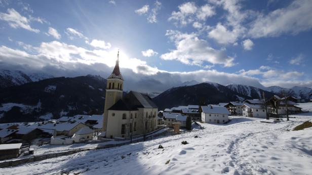 Eine verschneite Ortschaft mit Kirche vor einer Bergkulisse unter einem bewölkten Himmel.