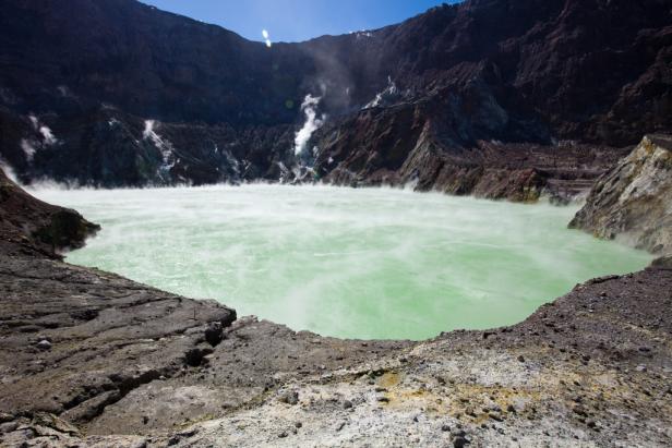 Ein trüber, grünlicher See in einem vulkanischen Krater mit aufsteigendem Dampf.