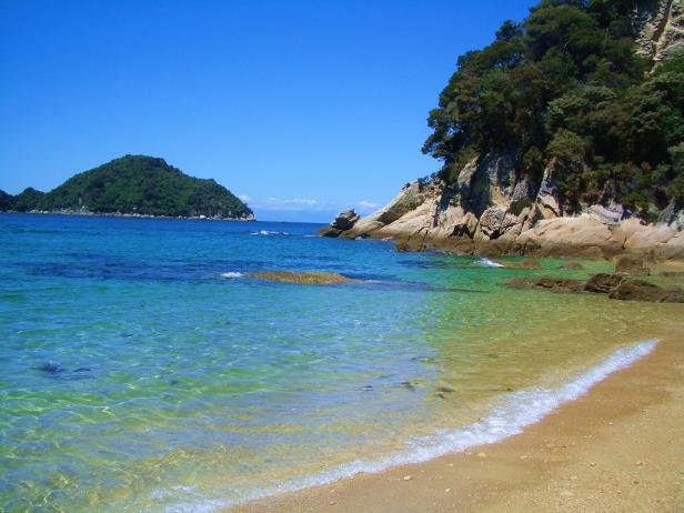 Ein Strand mit klarem, türkisfarbenem Wasser und einer kleinen, bewaldeten Insel im Hintergrund.