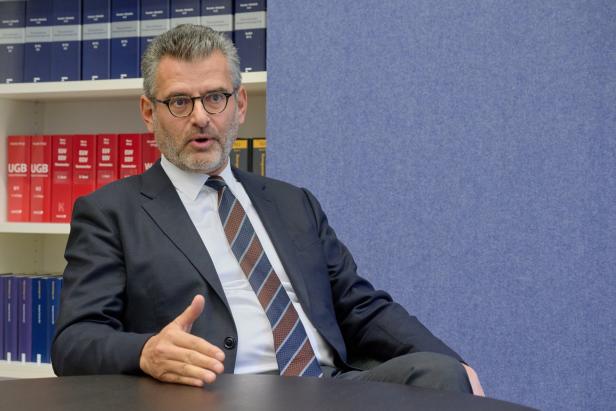 Ein Mann mit Brille und Anzug gestikuliert vor einem Bücherregal.