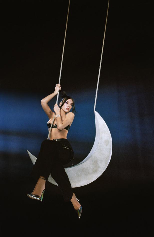 Dua Lipa sitzt auf einer Mondsichel-Schaukel vor einem dunklen Hintergrund.