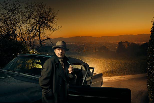 Ein Mann mit Hut und Mantel steht an einem Auto mit Blick auf eine Stadt bei Sonnenuntergang.