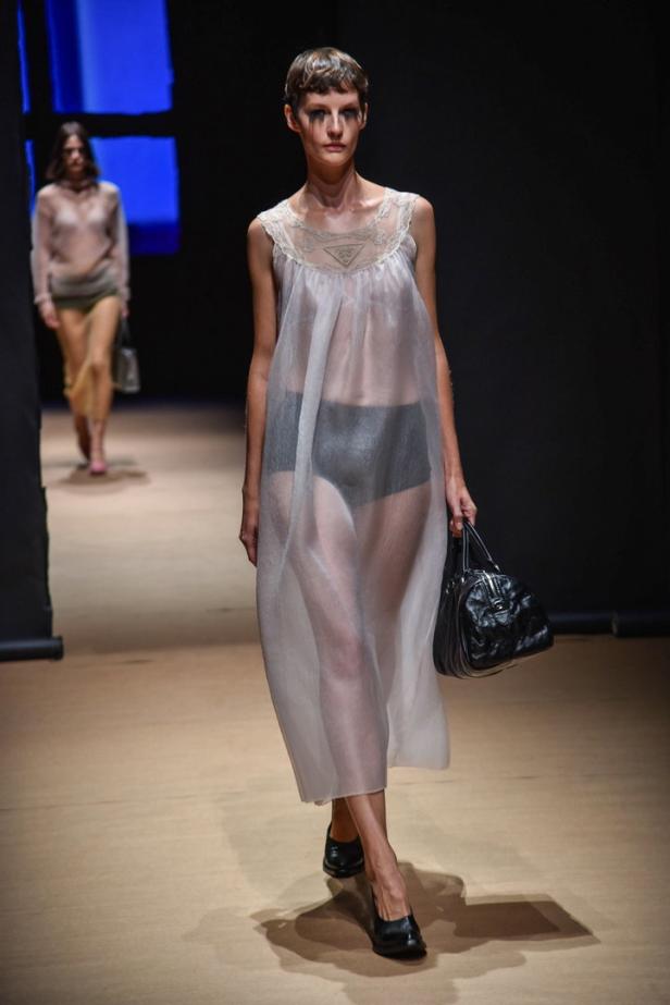Eine Frau in einem transparenten Kleid mit schwarzer Handtasche auf einem Laufsteg.