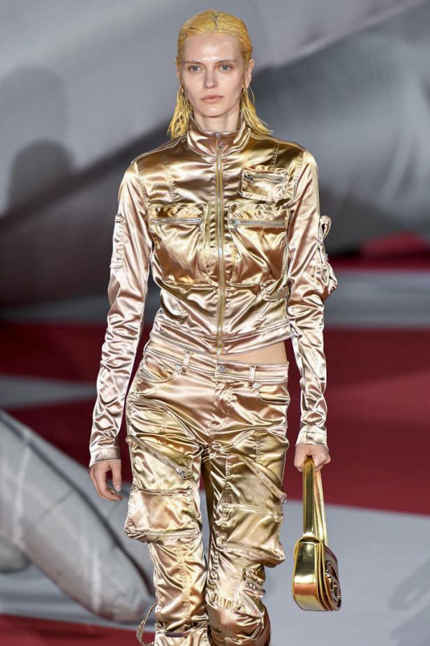 Ein Model präsentiert ein glänzendes, goldenes Hosen- und Jacken-Ensemble mit passender Handtasche.