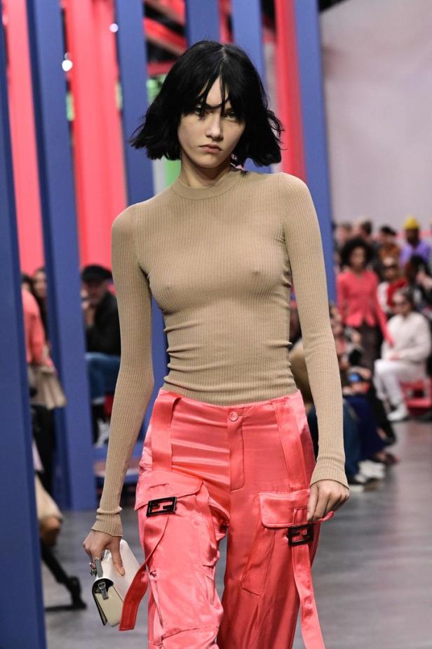 Ein Model mit beigem Oberteil und korallfarbenen Hosen auf einem Laufsteg.