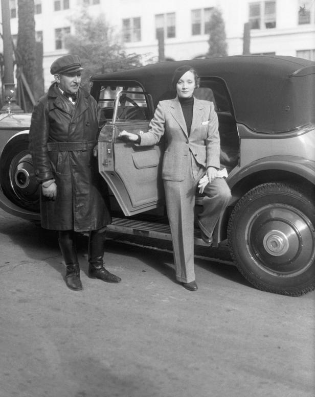 Marlene Dietrich steigt in einem Hosenanzug aus einem Auto, während ein Chauffeur daneben steht.