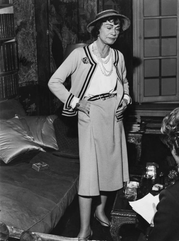 Coco Chanel posiert in einem Kostüm mit Blazer, Rock, Hut und Perlenkette.