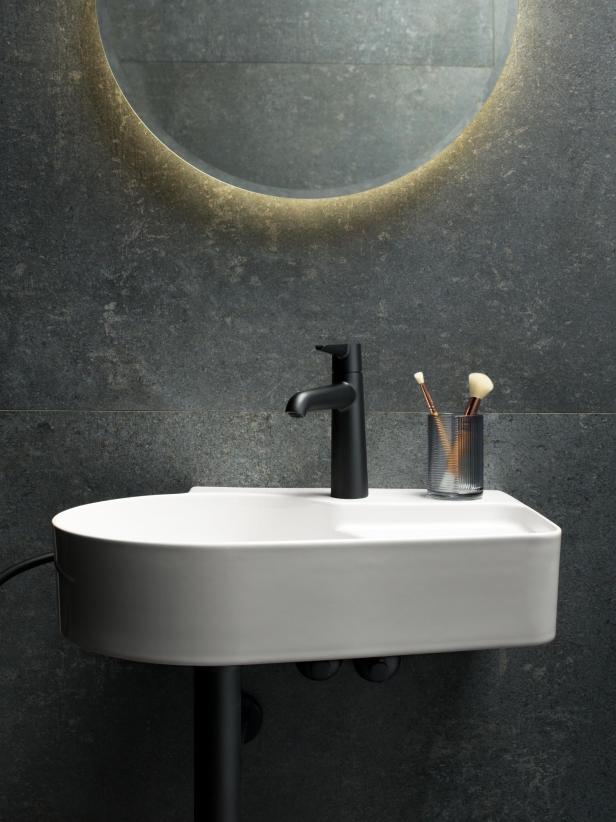 Ein Waschbecken mit schwarzer Armatur und zwei Schminkpinseln vor einer grauen Wand mit Spiegel.