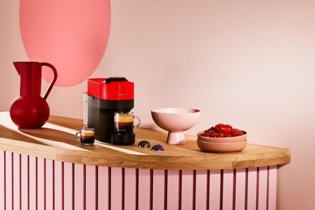Eine rote Nespresso-Maschine steht auf einem Tisch mit Tassen, Kapseln und einer Schale Tomaten.