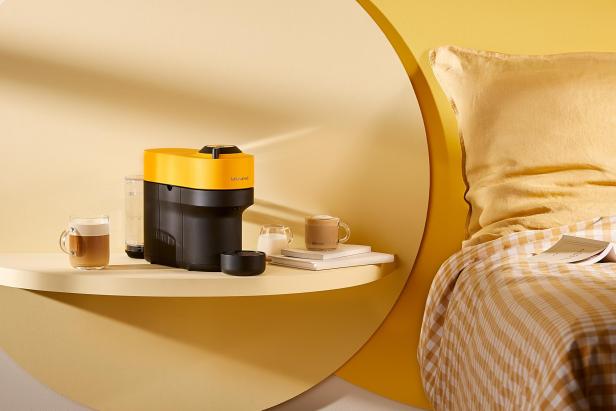 Eine gelbe Nespresso-Maschine steht mit Tassen auf einem Regal neben einem Bett.