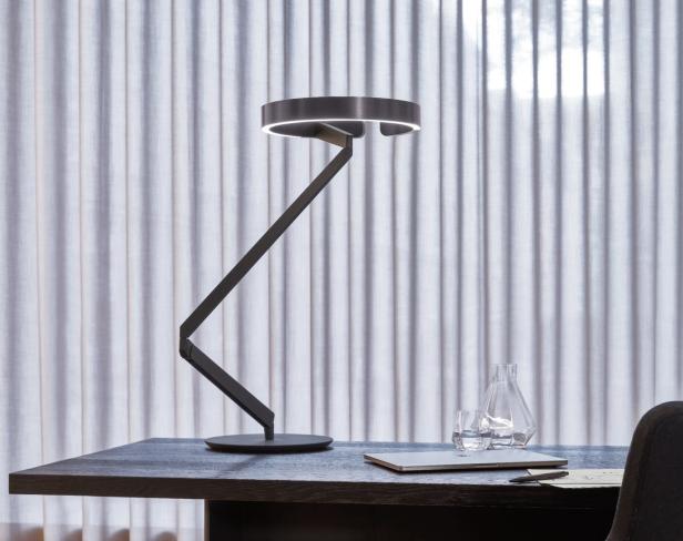 Eine moderne Schreibtischlampe beleuchtet einen Laptop und eine Wasserflasche auf einem dunklen Tisch.