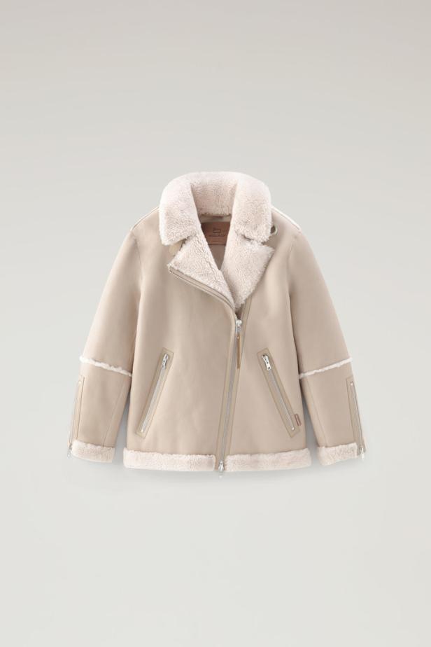 Eine hellbeige Shearling-Jacke mit silbernen Reißverschlüssen vor einem neutralen Hintergrund.
