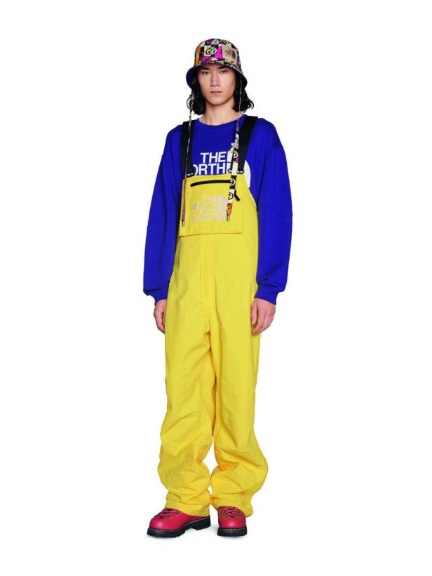 Ein Model trägt einen gelben Overall, einen lila Pullover und einen gemusterten Hut von The North Face x Gucci.