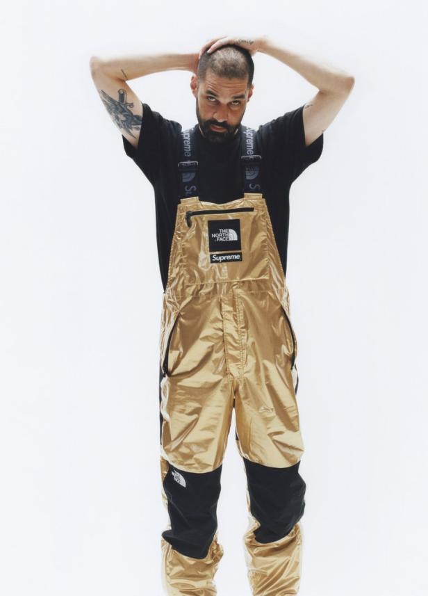 Ein Mann posiert in einer goldenen Latzhose von The North Face und Supreme.