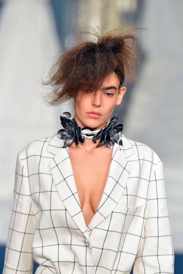 Eine Frau mit extravagantem Haar und einem karierten Blazer mit Blumenschmuck am Hals.