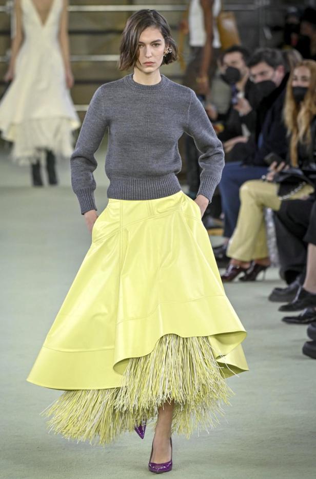 Ein Model präsentiert einen grauen Pullover und einen gelben Rock mit Fransen auf dem Laufsteg.