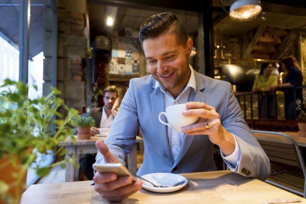 Ein Mann im Anzug sitzt in einem Café, hält eine Tasse Kaffee und schaut auf sein Smartphone.