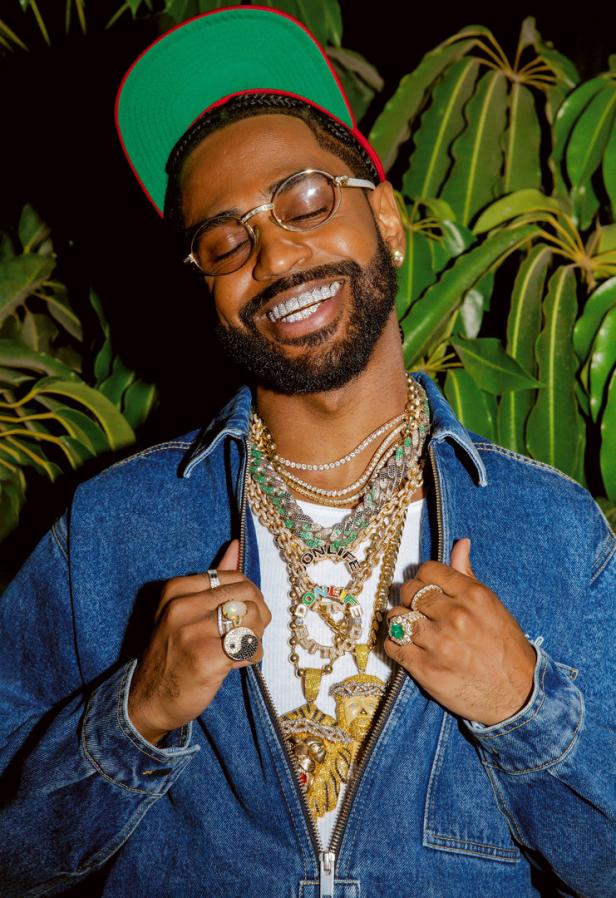 Big Sean mit Grillz und zahlreichen goldenen Halsketten vor tropischen Pflanzen.