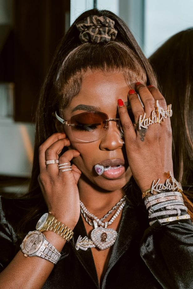Eine Frau mit Sonnenbrille und viel Schmuck, darunter Ringe mit der Aufschrift „Kash Doll“.