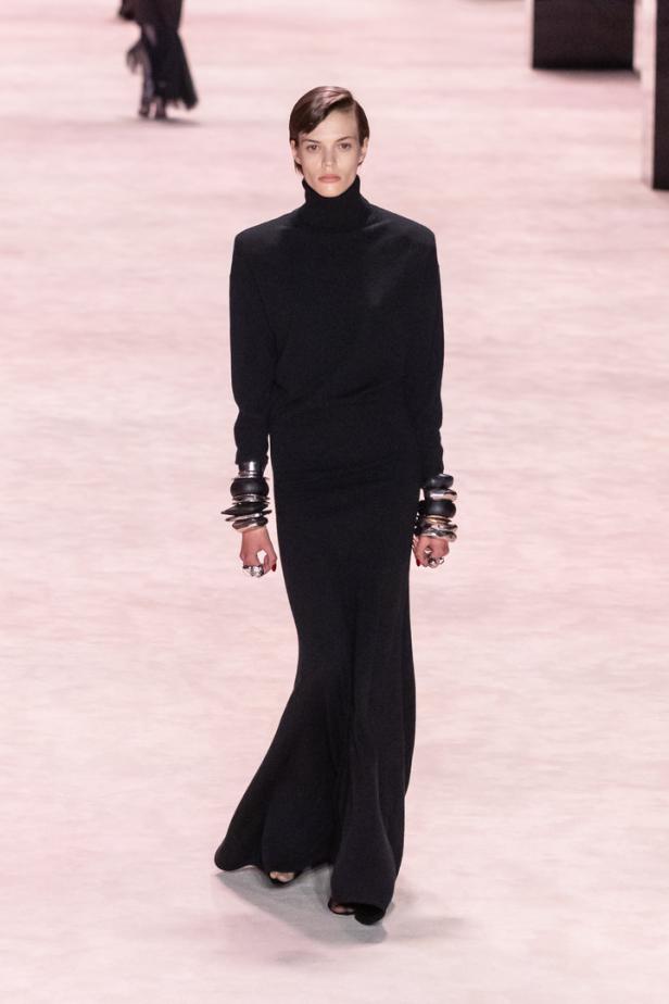 Ein Model in einem schwarzen Rollkragenkleid mit auffälligen Armreifen auf einem Laufsteg.