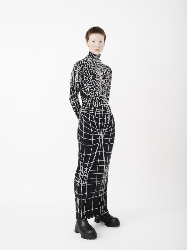 Eine Frau in einem schwarzen Rollkragenkleid mit weißem Gittermuster und schwarzen Stiefeln.