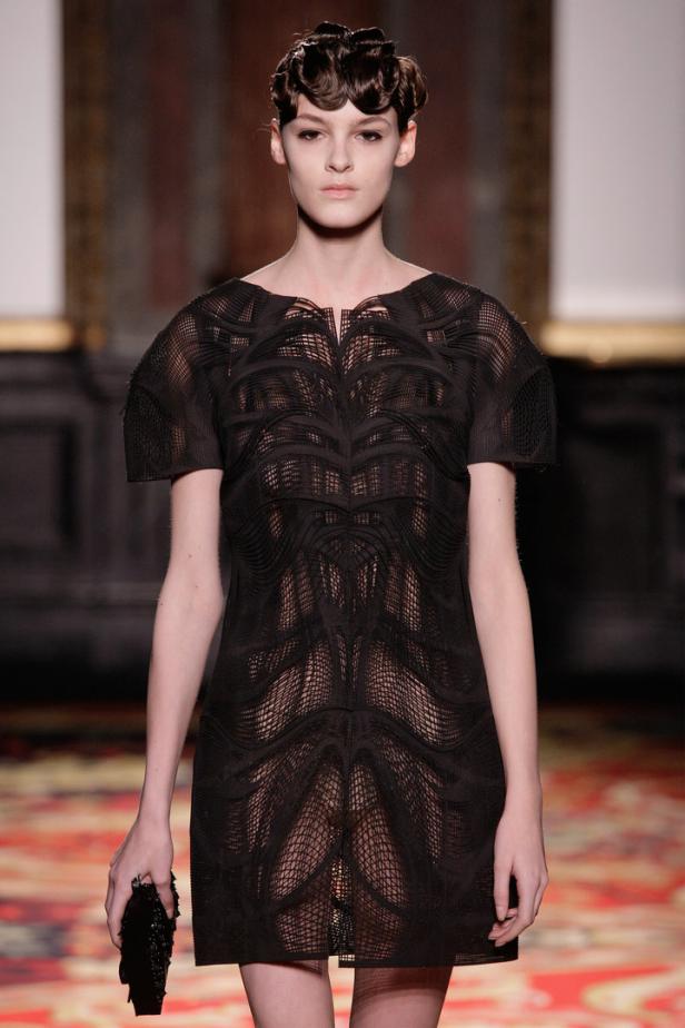 Eine Frau präsentiert ein schwarzes, durchscheinendes Kleid mit geometrischem Muster auf dem Laufsteg.