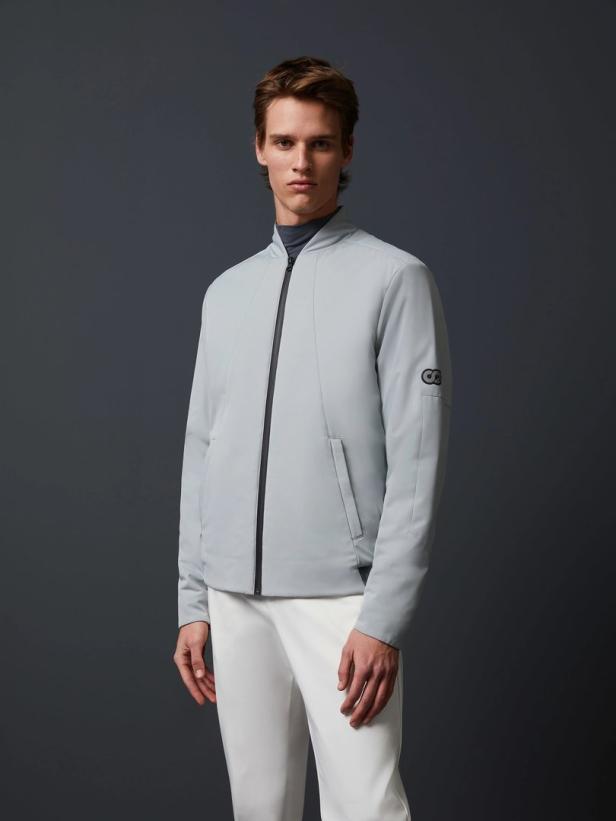Ein Mann trägt eine hellblaue Jacke und weiße Hose vor einem dunklen Hintergrund.