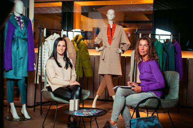 Zwei Frauen sitzen in einem Bekleidungsgeschäft zwischen Kleiderständern und Schaufensterpuppen.