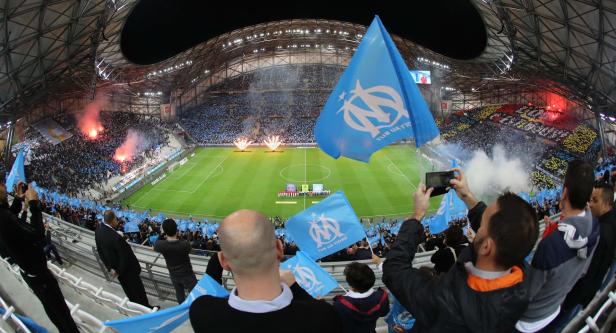 Ein Fußballstadion voller Fans, die blaue Fahnen schwenken und Feuerwerkskörper zünden.