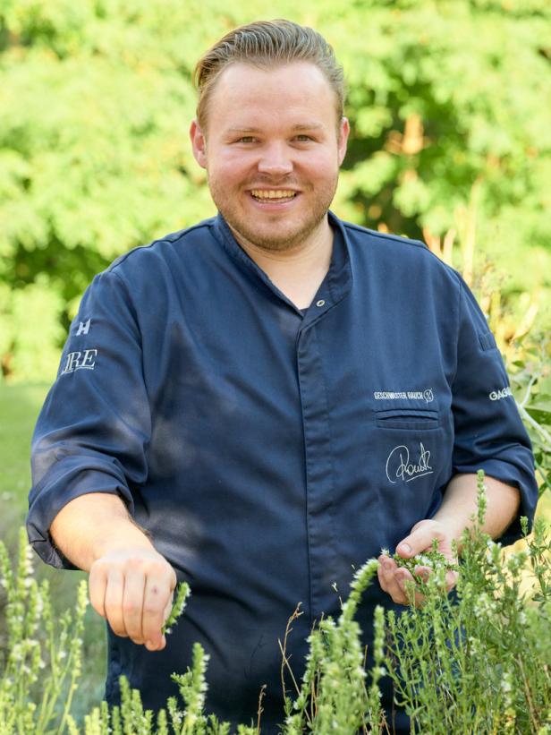 Ein lächelnder Koch in Kochjacke pflückt Kräuter im Garten.