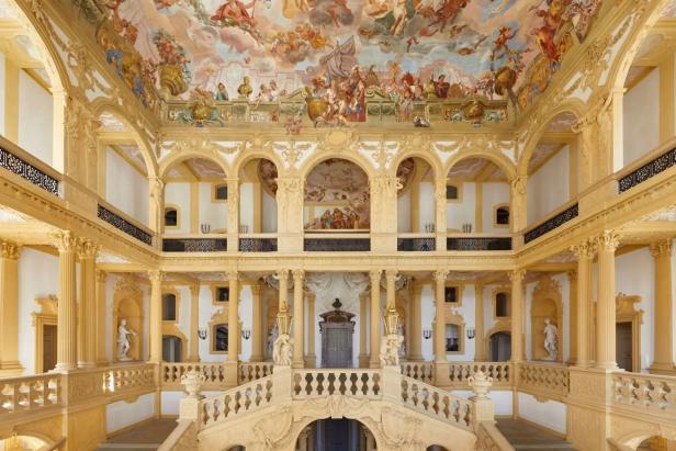 Eine prunkvolle Treppe führt in einen Saal mit Deckenmalerei und Säulen.