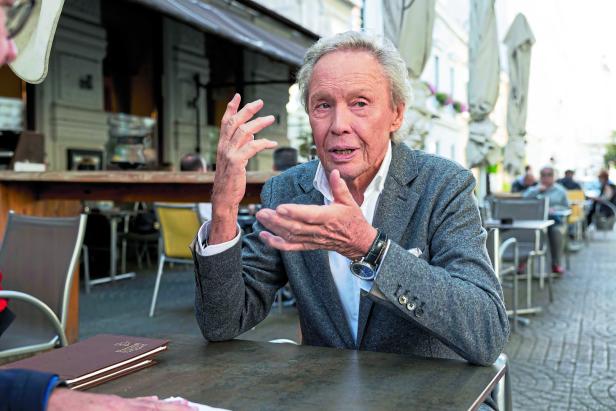 Ein älterer Mann gestikuliert an einem Tisch in einem Straßencafé.