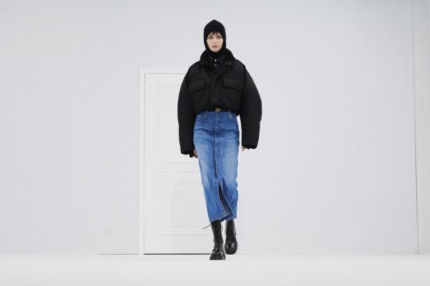 Eine Frau in Mütze, kurzer Jacke und Jeansrock posiert vor einer weißen Tür.