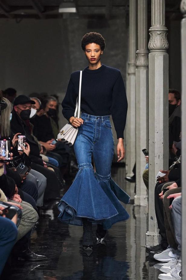 Eine Frau präsentiert eine Jeans mit ausgestelltem Saum auf einem Laufsteg.