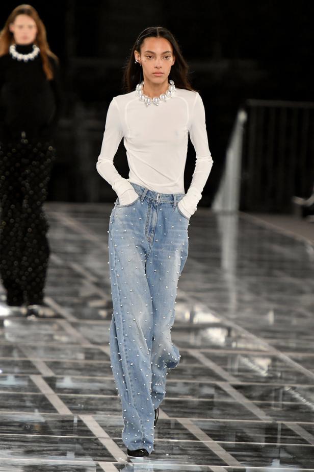 Eine Frau mit Perlenkette und Jeans mit Strasssteinen läuft auf einem Laufsteg.