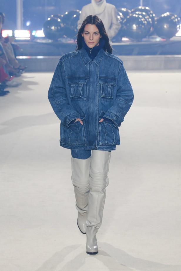 Eine Frau in einer übergroßen Jeansjacke und hohen Stiefeln läuft auf einem Laufsteg.