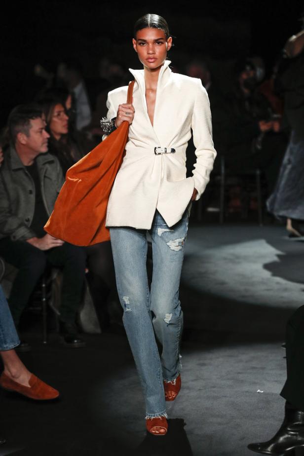 Ein Model auf dem Laufsteg mit einem weißen Blazer, Jeans und einer orangefarbenen Tasche.