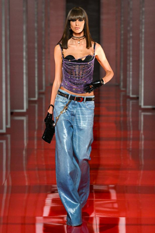 Ein Model auf dem Laufsteg mit einem lila Oberteil und glänzenden, weiten Jeans.