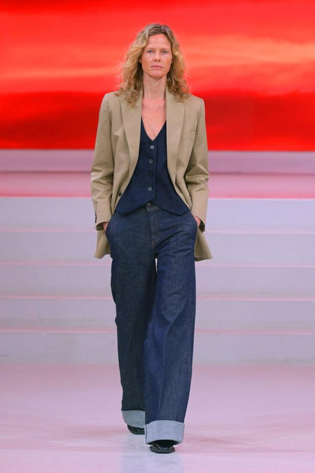 Eine Frau in einem beigen Blazer, einer Weste und Jeans posiert auf einem Laufsteg.