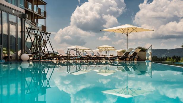 Ein Schwimmbad mit Liegestühlen und Sonnenschirmen vor einem Hotel unter blauem Himmel mit Wolken.
