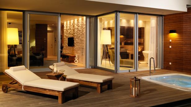 Zwei Liegestühle stehen auf einer Holzterrasse mit Whirlpool und Blick auf ein modernes Interieur.