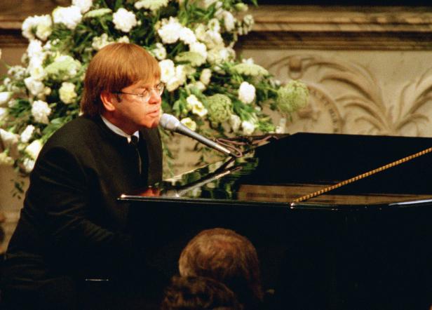 Elton John spielt Klavier und singt vor einem Blumenarrangement.