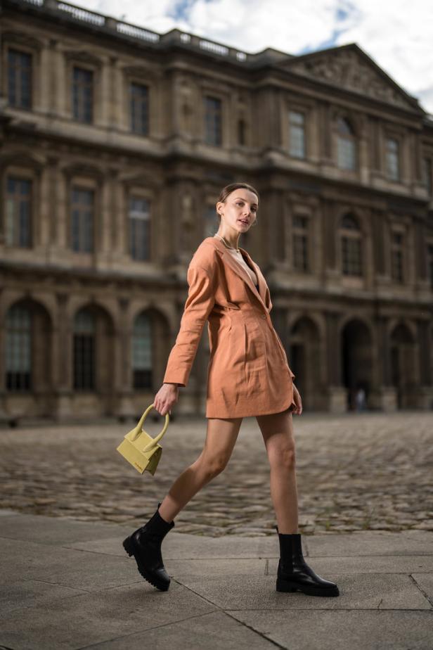 Eine Frau in einem orangefarbenen Blazer-Kleid und schwarzen Stiefeln geht mit einer gelben Handtasche vor einem Gebäude umher.