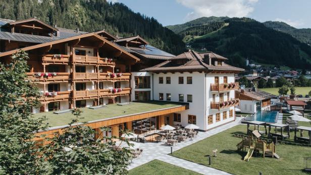 Ein großes Hotel im alpinen Stil mit Pool und Spielplatz vor einer bewaldeten Bergkulisse.