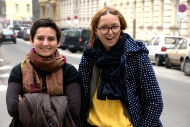 Zwei junge Frauen stehen lächelnd auf einem Bürgersteig in der Stadt.