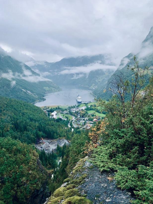 Blick über den Geirangerfjord in Norwegen mit einem Kreuzfahrtschiff und bewaldeten Bergen im Nebel.