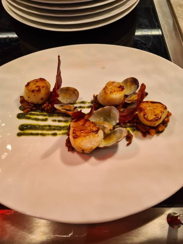Ein Teller mit Jakobsmuscheln, Venusmuscheln und Speck, garniert mit Pesto.