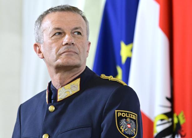 Ein älterer Mann in österreichischer Polizeiuniform steht vor EU- und österreichischer Flagge und blickt ernst zur Seite.
