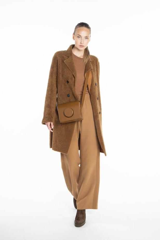 Eine Frau trägt einen braunen Mantel, eine passende Hose und eine runde Handtasche vor einem weißen Hintergrund.