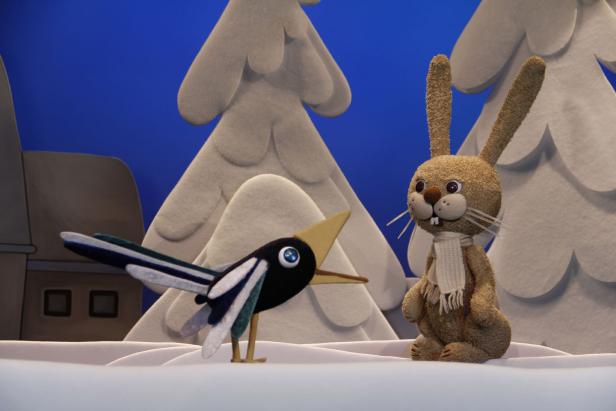 Hase Hoppla, Lilarum Ein Stoffhase mit Schal steht neben einer Stoffelster im Winterwald.